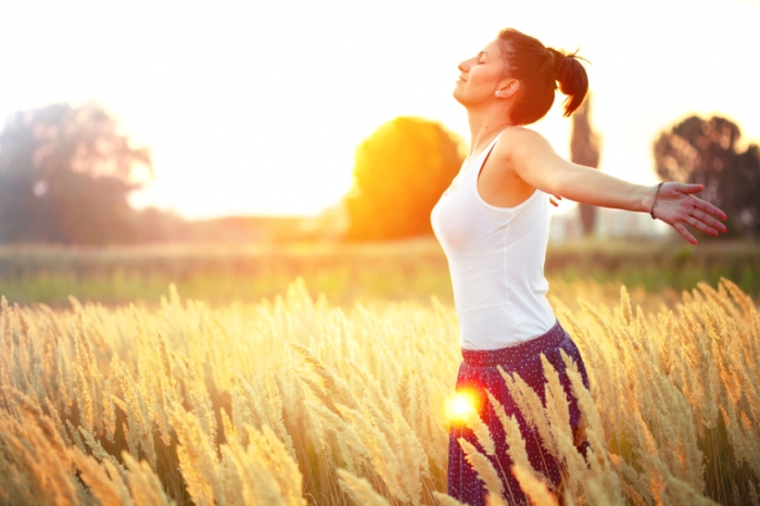 A woman wearing a white tank top stands with arms stretched wide towards her back as she stands basking in the golden glow of the sun in a field of wheat. 